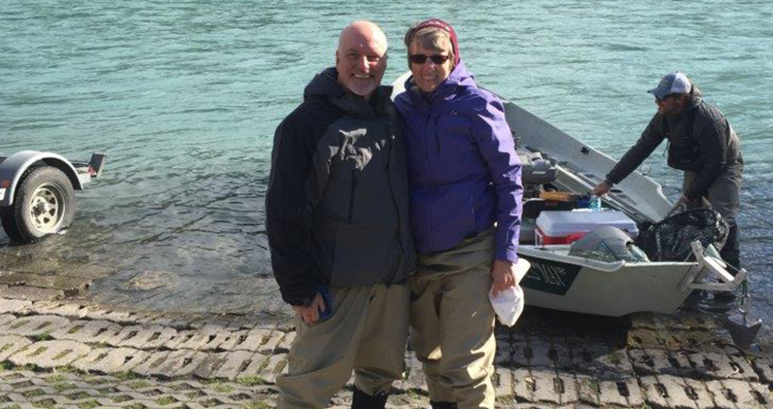 Two people in fishing gear smiling by a lake, with a boat and another person in the background.